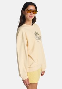 Εκρού φούτερ με γραφικό σχέδιο φοίνικα, στρογγυλή λαιμόκοψη και ριμπ μανσέτες, σε συνδυασμό με υφασμένα κίτρινα σορτς. Διάφανα κίτρινα γυαλιά ηλίου.