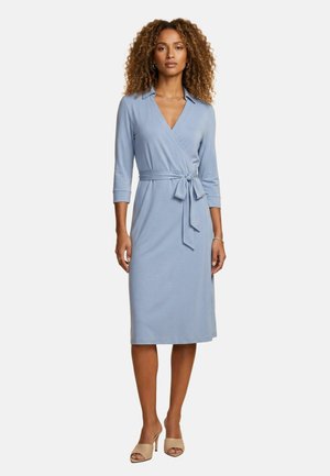 Femme portant une robe portefeuille bleu clair, longueur genou, à manches 3/4 et ceinture à la taille, debout en escarpins beiges sur un fond uni.