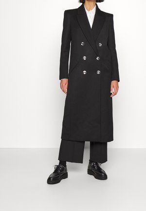 Osoba nosząca długi czarny dwurzędowy płaszcz z srebrnymi guzikami, czarne skrócone spodnie, czarne skarpety i czarne sznurowane buty.