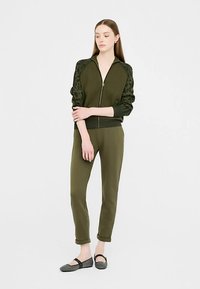 Olive green zip-up jacket with leopard pattern sleeves, paired with matching tapered pants. The model wears black flats and stands against a white background.