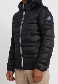 Czarna kurtka puchowa z kapturem, z kontrastowym białym i niebieskim zamkiem b�łyskawicznym, pikowanym wzorem oraz naszywką na lewym ramieniu.