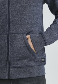 Felpa con zip color navy heathered, dotata di tasche laterali, polsini a coste e un piccolo marchio etichetta sull'orlo. Realizzata in tessuto leggero e testurizzato.