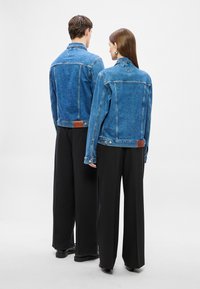 Kék farmerdzsekik klasszikus kialakítással, gombos mandzsettával és barna bőr címkével a hátoldalon, fekete, bő szárú nadrággal párosítva.