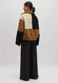 Fleecejacke mit einem Patchwork-Design in Braun, Creme und Schwarz. Sie hat einen kragenförmigen Hals und einen großzügigen Schnitt, kombiniert mit weiten schwarzen Hosen.