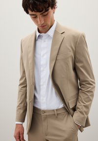 Uomo che indossa una giacca e pantaloni di velluto beige con una camicia bianca con bottoni, che guarda in basso con una mano in tasca.