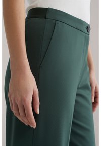 Krupan plan osobe u tamnozelenim krojenim hlačama s bočnim džepovima i kopčanim pojasom, uparenim s bijelom majicom.