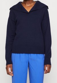 Navy blue knitted sweater with a wide folded collar, ribbed hem, and drop shoulders, paired with light blue loose-fitting trousers.
