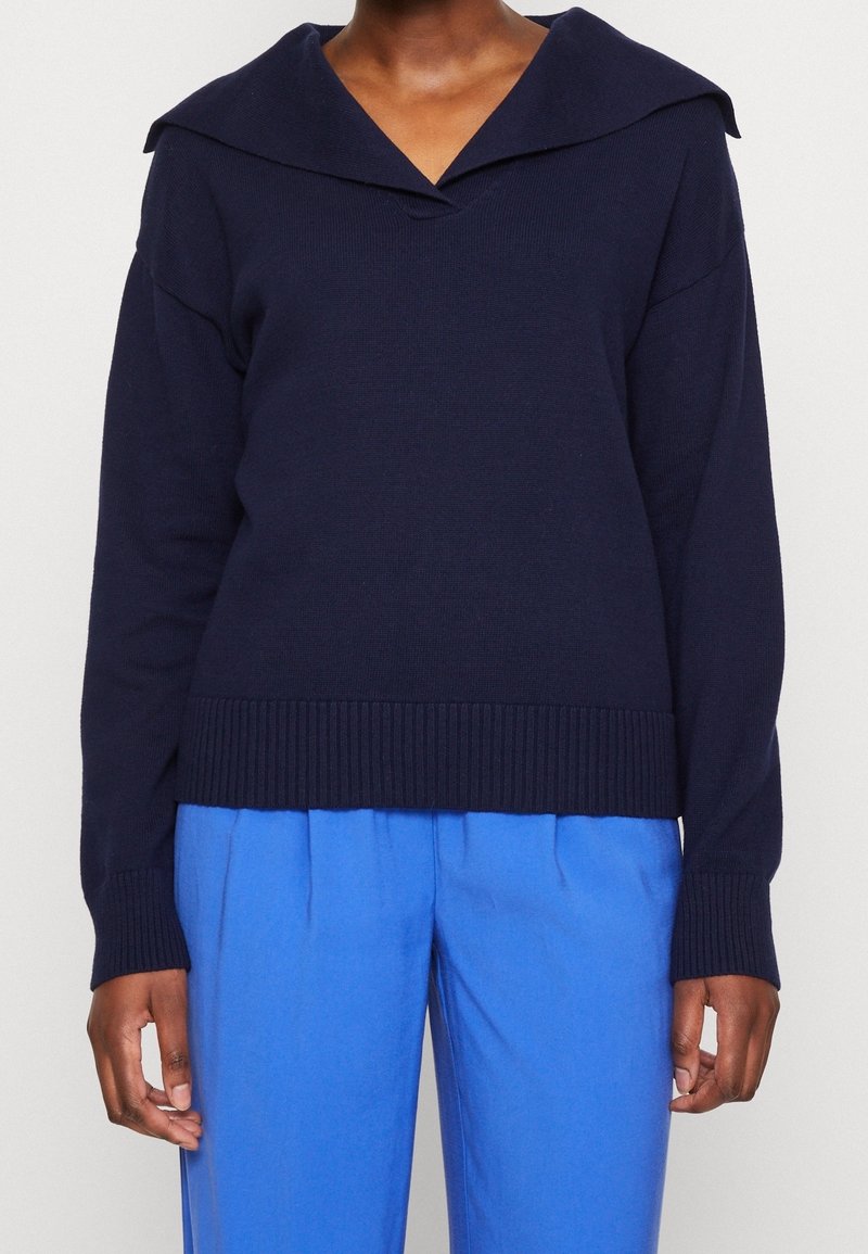Navy blue knitted sweater with a wide folded collar, ribbed hem, and drop shoulders, paired with light blue loose-fitting trousers.