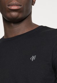 Zwarte katoenen t-shirt met een ronde halslijn en een klein grijs geborduurd logo op de linkerkant van de borst. Soepele textuur met een getailleerde vorm.