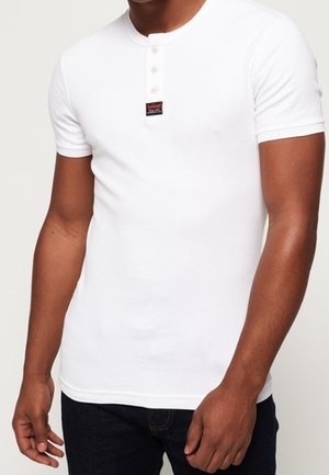 T-shirt henley blanc à manches courtes avec un col à boutonnière et des boutons clairs, fabriqué en tissu de coton, porté avec un jean foncé.