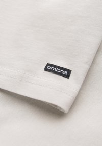 Hellgraues Baumwoll-T-Shirt mit glatter Textur, das ein schwarzes Etikett mit der Aufschrift "ombre" an der Ärmeln und doppelt genähte Nähte aufweist.