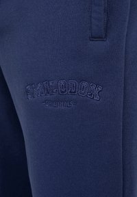 Marineblaue Sweatpants aus weichem Stoff, mit eingeprägten "SMILODOX" und "ORIGINALS" Logos am linken Oberschenkel.