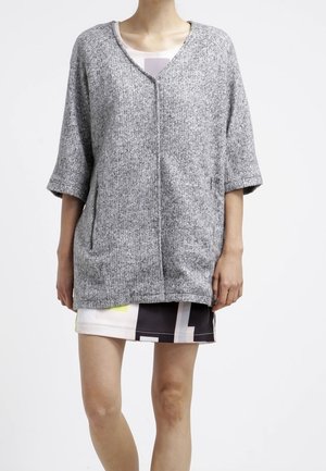 Osoba nosząca luźny szary kardigan z rękawami 3/4, na krótkiej spódnicy w geometryczne czarne, białe i żółte wzory.