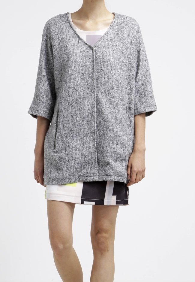 Personne portant un cardigan gris ample à manches 3/4 par-dessus une jupe courte à motifs géométriques noirs, blancs et jaunes.