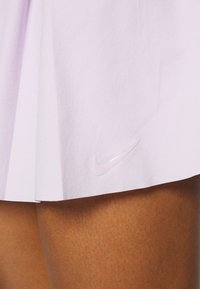 Falda plisada de color púrpura claro hecha de un tejido suave, con un logo de Nike en relieve y brillante en el dobladillo. La piel expuesta muestra un tono bronceado.