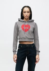 Šedá krátka mikina s kapucňou s červeným srdcom a logom, plus modré rifle s kontrastným šitím a ozdobným vreckom.