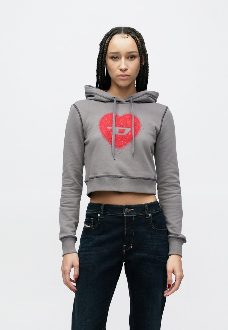 Šedá krátka mikina s kapucňou s červeným srdcom a logom, plus modré rifle s kontrastným šitím a ozdobným vreckom.