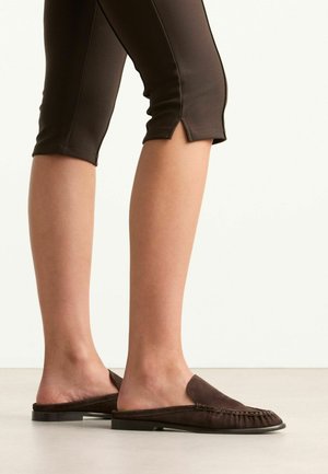 Jambes portant un pantalon court marron foncé et des mocassins marron foncé sans talons, debout sur un sol clair devant un fond uni.