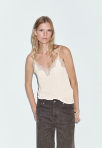 Camisole beige clair avec dentelle, présentant des fines bretelles, un design superposé et une texture douce, associé à un jean taille haute gris foncé.