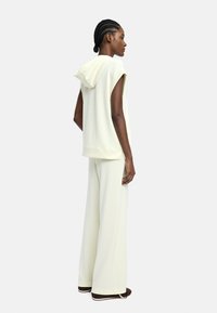 Kobieta z warkoczami, ubrana w bezrękawnik w kolorze kremowym i szerokie spodnie kremowe, stoi w profilu z rękami swobodnie opuszczonymi wzdłuż ciała.