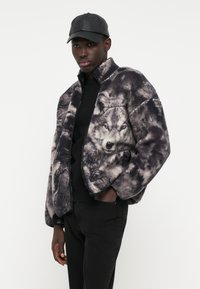 Ζακέτα από ψεύτικη γούνα με γραφικό σχέδιο λύκου, κλείσιμο με φερμουάρ, μαύρες λεπτομέρειες και άνετη εφαρμογή, συνδυασμένη με μαύρα τζιν και δερμάτινο καπέλο.