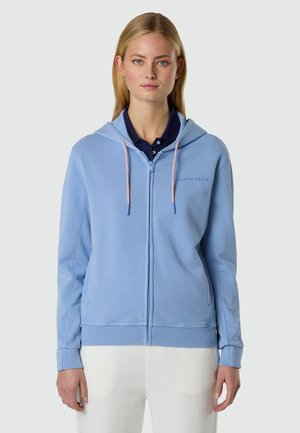 Donna che indossa una felpa leggera con zip azzurro chiaro e cordini rosa, con una polo blu navy sotto, in piedi davanti a uno sfondo semplice.