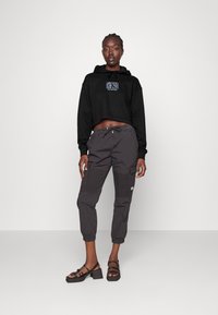 Hoodie nero cropped con logo grafico, abbinato a pantaloni cargo grigi scuri con tasche laterali e sandali neri con tacco a blocco e cinturini.