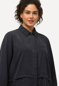 Giacca nera in tessuto lucido, con chiusura a bottoni, colletto e tasca sul petto, dal fit rilassato e oversize.
