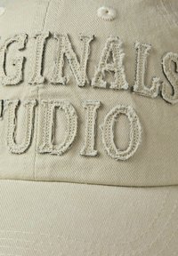 Bežna bejzbol kapa sa izdevenim, izvezenim tekstom "ORIGINALS STUDIO" u beloj i tamno sivoj boji, sa vidljivim šavovima i teksturiranim materijalom.