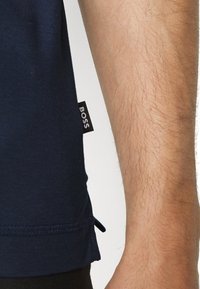 T-shirt bleu foncé avec une étiquette affichant "BOSS" sur le côté. Manches courtes avec une texture lisse et un ourlet légèrement courbé.