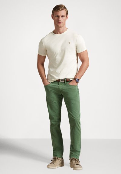 T-shirt en coton crème, pantalon vert et baskets beige. Comprend un petit logo sur le t-shirt, une ceinture et une montre avec un cadran bleu et des accents métalliques.
