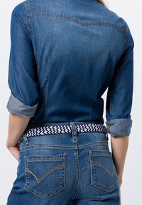 Chemise en denim aux manches retroussées avec un empiècement dos, associée à un jean bleu ceinturé d'une ceinture tissée à motifs en bleu marine et blanc.