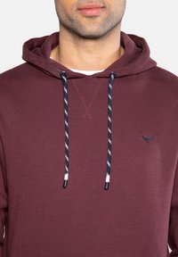 Bordeauxkleurige hoodie met een trekkoordcapuchon, voorzien van marineblauwe en witte gestreepte koorden, en een klein geborduurd logo op de borst. Zacht aanvoelend.