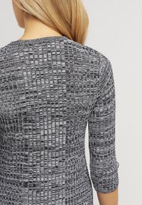 T-shirt tricoté texturé gris et noir avec des manches trois-quarts, présentant un motif de grille et un col rond. Le tissu semble doux et extensible.