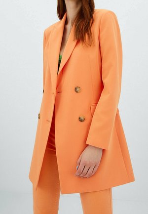 Femme portant un blazer orange croisé avec un pantalon assorti et un haut vert, posant devant un fond clair.
