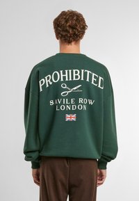 Zelená mikina s nápisom "ZAKAZANÉ" v veľkom bielom písme a grafikou nožníc. Pod ním sa nachádza nápis "SAVILE ROW LONDON" a akcent britskej vlajky.