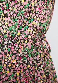 Gros plan d’un tissu à imprimé floral dense en rose, jaune, vert et blanc sur fond noir, avec un détail de nœud noué sur un vêtement.