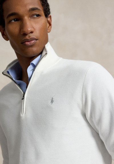 Homme portant un pull texturé blanc à fermeture éclair au quart, par-dessus une chemise bleu clair à col, regardant sur le côté sur un fond neutre.