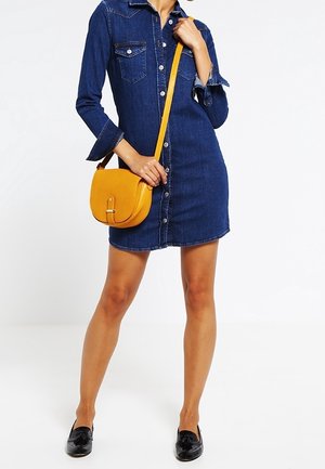Femme portant une robe-chemise en denim boutonnée avec des manches retroussées, portant un sac bandoulière jaune et des mocassins noirs.