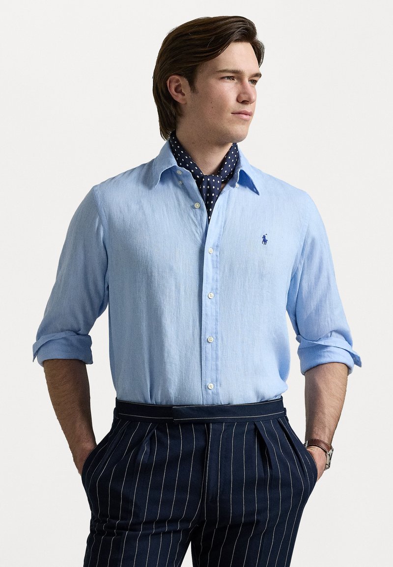 Világoskék vászon ing gombos kivitelben, feltekeredett hosszú ujjakkal. Tengerészkék csíkos nadrággal és pöttyös sállal párosítva.