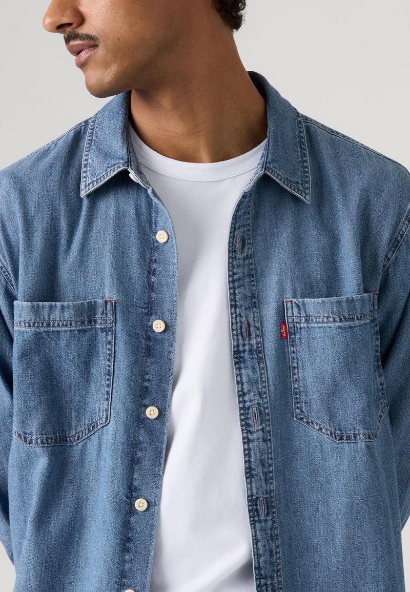 Homme portant une chemise en jean bleue boutonnée sur un t-shirt blanc, regardant sur le côté, avec une moustache taillée et des cheveux bouclés foncés.