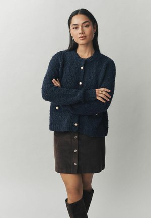 Marineblauer, strukturierter Cardigan mit Rundhalsausschnitt und Knopfverschluss, kombiniert mit einem braunen Cordrock mit vertikaler Knopfleiste.
