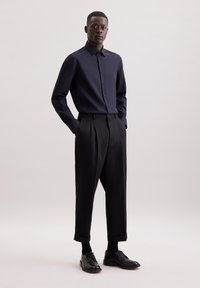 Camicia azzurra con bottoni e maniche lunghe, abbinata a pantaloni neri a pieghe. Le scarpe sono in pelle nera con una finitura liscia.