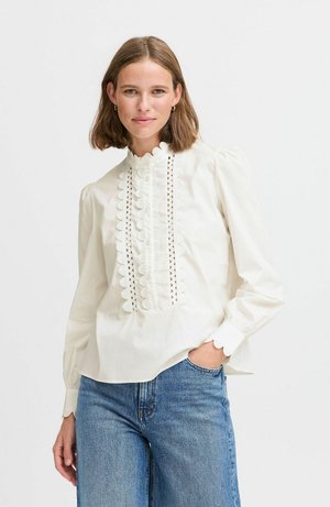 Blusa bianca con colletto alto arricciato, dettagli in pizzo verticali e maniche a sbuffo, abbinata a jeans di denim azzurri.