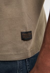 Olijfgroene katoenen T-shirt met een zwart label aan de zoom, met witte en oranje tekst en een gestikte naaddetail.