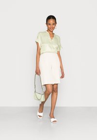 Blusa verde-clara de mangas curtas com decote em V, combinada com calções bege. Os acessórios incluem uma pequena mala verde-clara e sandálias brancas.