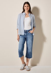Světle modrá a bílá pruhovaná košile na knoflíky přes bílý top bez rukávů, doplněná o zkrácené modré džíny a šedé tenisky. Hladké materiály a volný střih.