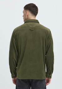 Groene fleecetop met lange mouwen en knoopmanschetten. Heeft een kraag en een lus om op te hangen. Zachte textuur en relaxte pasvorm.