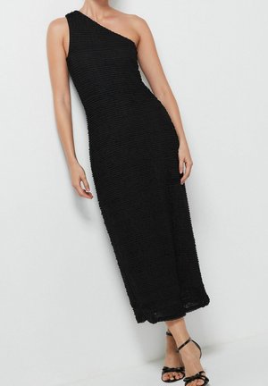 Kobieta ubrana w czarną, jednobarwną sukienkę na jedno ramię o fakturowanej tkaninie oraz czarne, paskowane sandały na wysokim obcasie, stojąca na tle białej, gładkiej ściany.