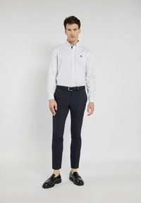 Biała koszula z guzikami w małe, ciemne kropki, granatowe spodnie w slim fit, teksturowany ciemny pasek i czarne loafersy. Ubranie jest casualowe, ale jednocześnie eleganckie.